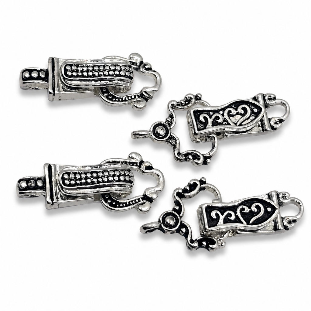 Silver-toned fold-over magnetic clasps with an ornate scroll and heart design are displayed on a white background. This jewelry finding set includes four matching clasp components shown from both front and back views. The decorative front features black enamel-style detailing with elegant swirls and heart accents, while the reverse side showcases textured beaded patterns. These secure magnetic clasps are ideal for bracelet making, bead weaving projects, necklaces, and handmade jewelry designs.