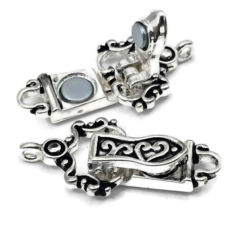 Decorative single-strand fold over magnetic clasp in bright silver finish featuring elegant black enamel scrollwork accents and ornamental detailing. Shown in both open and closed positions to demonstrate the hinged fold-over design and magnetic closure system. Each end includes a single attachment loop for connecting jewelry designs. Perfect for handmade bracelets, beading projects, artisan jewelry, magnetic bracelet closures, and custom jewelry making supplies.