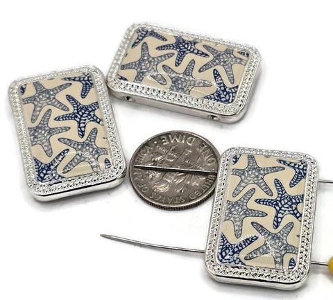 Three rectangular silver-toned pendants with a starfish pattern. The starfish are navy blue on a cream background. The pendants have a silver-toned beaded border. A dime is next to the pendants for size comparison. The pendants are on a white surface.