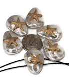 A collection of silver-colored, pebble-shaped beads, each adorned with a gold-colored starfish. The starfish are raised and detailed. A dime is placed among the beads for size comparison. A black cord is visible at the bottom, suggesting the beads are for jewelry making.