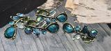 These botanical 2‑hole sliders feature deep teal glass rhinestones framed by beaded bezels and antique brass vines. Enamel leaves in sage and indigo add layered color. Dual threading ensures a flat, secure fit for bracelets or necklace focal bars. Ideal for garden, mermaid, or Art Nouveau jewelry that calls for luminous sparkle and intricate metalwork.