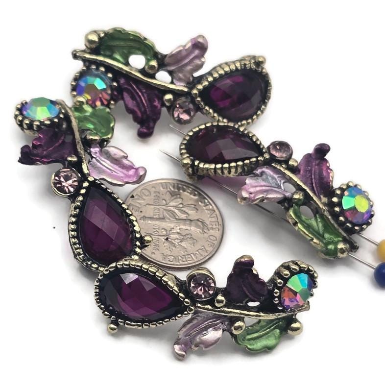 These botanical 2‑hole slider beads feature faceted resin teardrops in deep plum, accented with Austrian crystals and rhinestones. Antique brass/gold settings wind into leafy vines with hand‑tinted enamel leaves in lavender and green. Dual holes keep the focal flat for bracelets, chokers, or pendant bars. Perfect for boho luxe, cottagecore, and Art Nouveau jewelry needing romantic color and secure alignment.