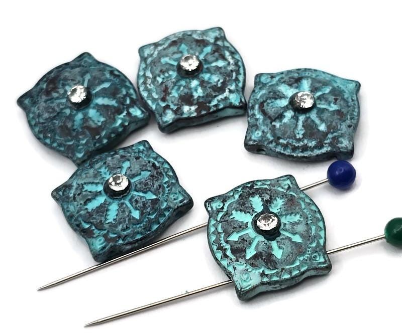 Turquoise patina 2 hole slider beads with a boho medallion motif and sparkling crystal center. These unique beads make instant focal beads for bracelets or pendants. Rich verdigris texture over dark metal adds depth, while the square pillow shape delivers sculpted dimension in photos. Premium metal findings with a durable, high‑quality finish. Great for artisan, vintage, festival, and Southwestern‑inspired jewelry collections.