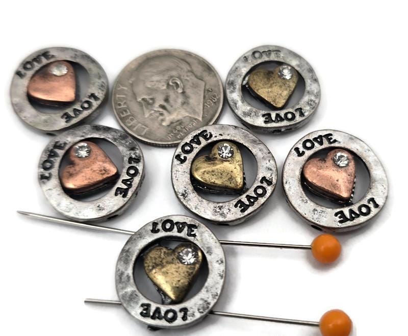 Add heartfelt charm to your creations with these metal 2‑hole “LOVE” heart slider beads. Each bead features a raised golden or copper heart surrounded by a ring engraved with the word “LOVE” and accented by a tiny crystal detail. Perfect as focal beads for bracelets, necklaces, or mixed‑metal designs. Durable, artistic, and easy to string through two holes for a secure, professional finish in your handmade jewelry.