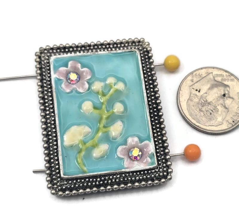 Make a statement with this unique focal 2-hole slider bead, featuring a serene aqua enamel background and a delicate floral design. Framed in textured antique silver metal, this large rectangular bead is perfect for creating high-end bracelets and necklaces. The two-hole construction ensures your jewelry stays flat and aligned. Pair with seed beads, pearls, or gemstone beads for a professional, artisan look in your handmade jewelry projects.