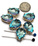 Aquamarine pear-shaped rhinestone slider beads displayed in a decorative product photograph with reflective surfaces and soft lighting. The faceted rhinestones reveal vibrant blue tones enhanced by flashes of violet, pink, turquoise, and lavender. Polished gold settings frame each stone while dual holes provide versatile stringing options. These sparkling beads are perfect for handmade jewelry, bead weaving, and statement bracelet designs.