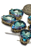 Pear-shaped Aquamarine rhinestone slider beads featuring two parallel stringing holes and polished gold settings. Displayed in close-up photography, the faceted stones reflect shades of aquamarine blue with flashes of pink, violet, lavender, and turquoise. Their elegant shape and sparkling finish make these 2 Hole Slider Beads ideal for handmade jewelry projects, bracelet focal designs, necklaces, and decorative beadwork.