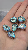 Six Aquamarine rhinestone slider beads arranged on a reflective black surface, creating dramatic reflections beneath each bead. The faceted pear-shaped stones display rich aquamarine blue color accented by subtle violet, lavender, pink, and turquoise highlights. Set in polished gold settings with two parallel stringing holes, these 2 Hole Slider Beads make beautiful focal pieces for bracelets, necklaces, earrings, and custom jewelry designs.
