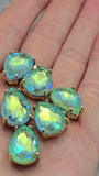 Pear-shaped Seafoam AB rhinestone beads with two parallel stringing holes displayed alongside a dime and beading needles. The faceted surface reflects vibrant shades of seafoam green, aqua blue, turquoise, and lime as light moves across the Aurora Borealis finish. Set in polished gold settings, these sparkling slider beads are designed for creating elegant bracelets, necklaces, earrings, pendants, and multi-strand jewelry designs with professional-looking results.