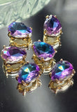 Gold pear-shaped 2 hole slider bead featuring a sparkling Purple Aqua AB rhinestone. The faceted stone reflects brilliant shades of amethyst purple, aqua blue, turquoise, and teal. These decorative focal beads are perfect for jewelry making, custom bracelets, necklaces, earrings, bead embroidery, loom work, and artisan beading projects.