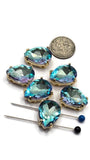 Close-up of Aquamarine rhinestone 2 Hole Slider Beads arranged beside a dime and jewelry needles. The faceted pear-shaped rhinestones display vibrant aquamarine blue coloring accented by subtle flashes of violet, pink, lavender, and turquoise. Each bead is set in a polished gold setting and features two parallel stringing holes, making them perfect for bracelet making, necklace designs, and bead weaving projects.