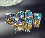 Sparkling Aquamarine pear-shaped slider beads with faceted rhinestones shown on a white background. The polished gold settings complement the brilliant blue stones, which display shifting flashes of violet, lavender, turquoise, and pink. A dime and beading needles provide scale. These 2 Hole Slider Beads are ideal for creating elegant bracelets, necklaces, earrings, pendants, and custom handmade jewelry with professional-looking results.