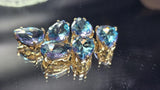 Aquamarine rhinestone 2 Hole Slider Beads displayed on a reflective black surface with soft lighting. The faceted pear-shaped stones showcase deep aquamarine blue tones accented with flashes of violet, lavender, turquoise, and pink. Their polished gold settings add warmth and contrast. These sparkling dual-hole beads are perfect focal components for handmade jewelry, bead weaving, bracelet making, and elegant necklace designs.