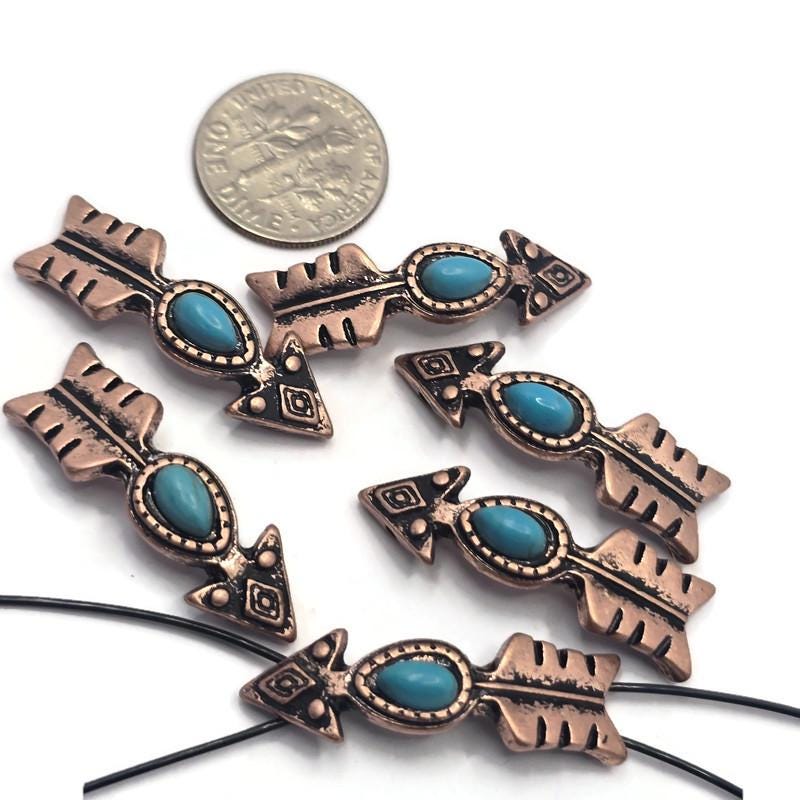 Arrow-shaped 2 Hole Slider Beads featuring faux turquoise center stones set in antique copper metal. Displayed with a dime for size reference, these western-inspired focal beads showcase decorative arrowhead details and feathered tails. Perfect for handmade bracelets, necklaces, leather jewelry, Southwestern accessories, artisan jewelry projects, and custom western-themed jewelry making designs.
