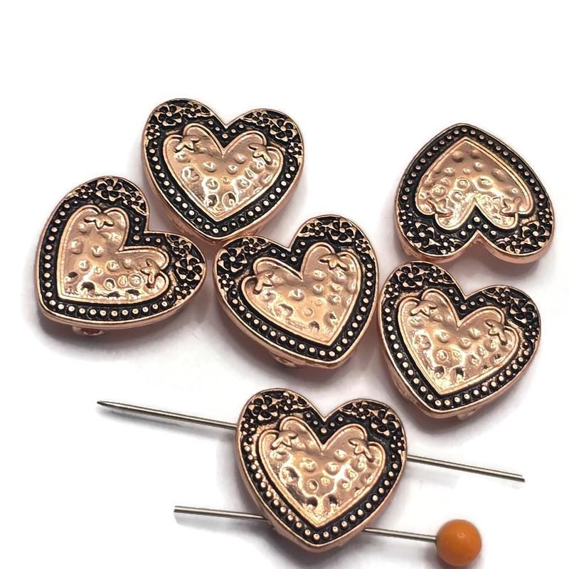 Add warmth and vintage charm to your handmade jewelry with these Heart 2 Hole Slider Beads. Featuring a bright copper finish with antique detailing, these decorative focal beads showcase a raised textured heart surrounded by ornate scrollwork. Perfect for bracelet making, necklace designs, earrings, pendants, and multi-strand jewelry, their dual-hole construction provides stability while adding romantic style to artisan jewelry and custom handmade creations.