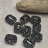 Add dramatic sparkle to handmade jewelry with these jet and hematite rhinestone slider beads in a sleek gunmetal finish. Each 2 hole bead features a faceted rectangular center crystal framed by smaller smoky rhinestones for added brilliance and dimension. Their dark neutral coloring makes them perfect for modern bracelets, gothic jewelry, evening designs, bead weaving, and necklaces. Pair them with crystals, gemstones, seed beads, chain, or metallic spacers for unique jewelry designs.