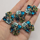 Blue floral 2 hole slider beads in an antique gold finish featuring vivid Blue Zircon rhinestones, blue enamel flowers, and teal green leaves. These statement focal beads are beautiful jewelry making supplies for double strand bracelets, bead weaving, necklaces, seed bead jewelry, boho designs, and romantic botanical accessories. Dimensional vines and sparkling crystals add texture and brilliant color.