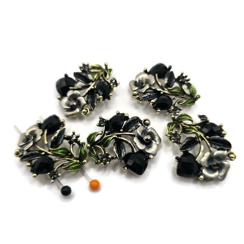 Antique gold floral 2 hole slider beads feature faceted jet black rhinestones, green and gray enamel accents, and beautifully detailed dimensional flowers and leaves. These dramatic focal beads are perfect for bracelets, necklaces, bead weaving, and multi-strand jewelry designs. The two-hole construction helps keep strands aligned while adding a professional finished look. Each bead measures approximately 30 mm x 21 mm with 12 mm between the loops. Five beads per package.