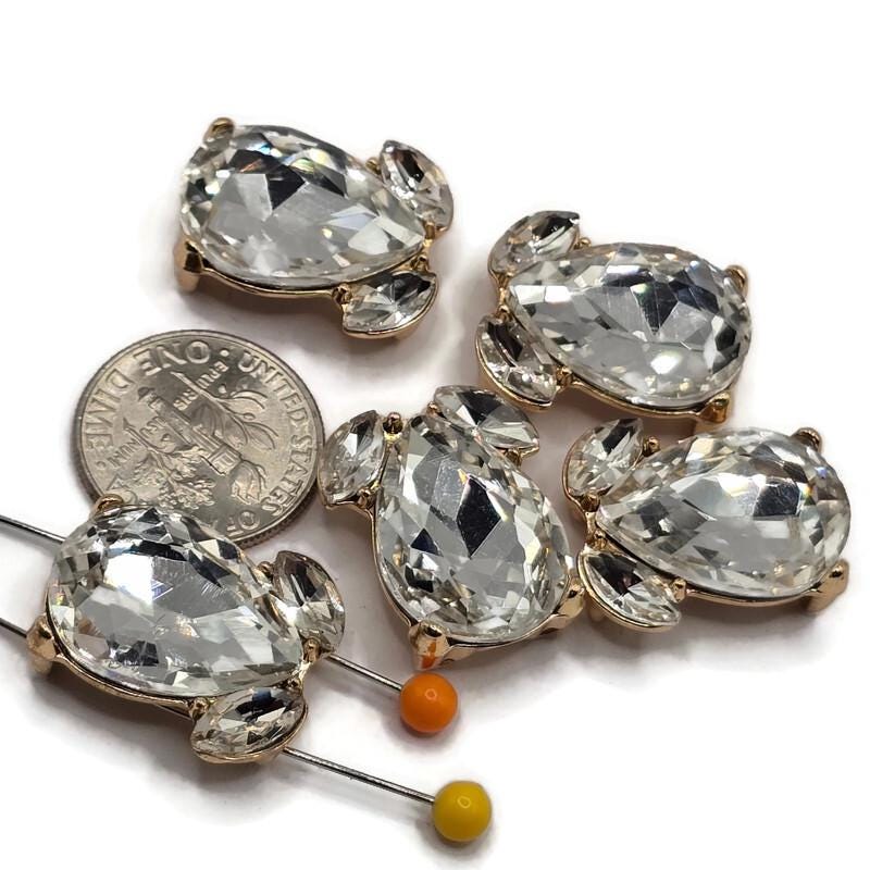 Clear rhinestone 2 hole slider beads featuring large faceted teardrop crystals with two smaller coordinating stones in a polished bright gold finish. Each 20 mm focal bead has two hidden stringing loops approximately 9 mm apart on the back. Sparkling jewelry components for double-strand bracelets, bead weaving, necklaces, bridal jewelry, statement designs, handmade accessories, and elegant jewelry-making projects.