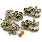 Green enamel flower 2 hole slider beads featuring a bright gold finish, dimensional floral design, and coordinating emerald, sage, and pearly ivory colors. These botanical focal beads are approximately 26 mm long and 17 mm wide with 7.5 mm between holes. Ideal jewelry-making components for double-strand bracelets, bead weaving, necklaces, garden-inspired jewelry, nature designs, and handmade artisan accessories.