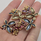 Beautiful gold floral focal beads featuring contrasting charcoal gray, burgundy, smoky champagne, purple, and navy blue enamel flowers. Each decorative jewelry component showcases three overlapping blossoms with elongated textured petals, polished gold borders, and delicate openwork metallic flower accents. These 2 Hole Slider Beads are perfect for handmade jewelry designs, multi-strand bracelets, necklace making, and bead weaving.