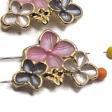 Detailed view of bright gold floral slider beads featuring three overlapping enamel flowers in complementary pink, gray, and pearl white colors. The larger pink flower showcases delicately textured petals, while the smaller smoky gray and pearl white flowers provide beautiful contrast. Polished gold outlines and raised centers highlight the dimensional botanical design. Perfect for handmade bracelets, necklaces, bead weaving, and romantic garden-inspired jewelry.