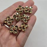 Detailed view of bright gold smoked topaz rhinestone 2 hole slider beads measuring approximately 14 mm wide and 14 mm tall, with 8 mm between the holes. Each decorative bead features four round golden-brown rhinestones in individual polished gold settings. The compact floral-inspired arrangement adds sparkle and dimensional detail to handmade jewelry and double-strand bracelet designs.