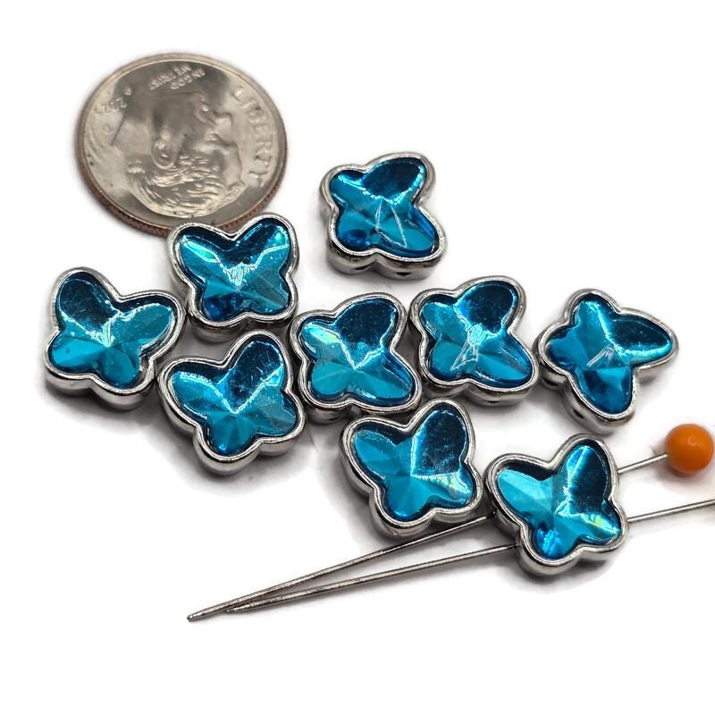 Nine electric blue butterfly 2 hole slider beads arranged on a white background. Each butterfly features a vibrant faceted blue stone with turquoise, aqua, and deep blue reflections surrounded by a polished platinum silver metal setting. These small decorative butterfly focal beads measure approximately 11 mm wide and 10.5 mm tall, perfect for handmade bracelets, necklaces, bead weaving, and multi-strand jewelry designs.