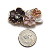Close-up of bright gold floral 2 hole slider beads featuring a large charcoal-black enamel flower with a sparkling clear rhinestone center. Smaller soft pink and peach enamel flowers surround the dark bloom, accompanied by delicate pearl white leaves. Polished gold outlines and raised flower centers highlight the dimensional floral arrangement, creating elegant decorative focal beads for handmade jewelry.