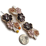 Bright gold floral 2 hole slider bead threaded onto two jewelry-making pins to demonstrate the side-drilled holes and approximately 8 mm strand spacing. The dimensional focal bead features a charcoal-black flower with a clear rhinestone center, smaller pink and peach enamel flowers, and pearlescent white leaves. Decorative multi-strand jewelry component for double-strand bracelets, necklaces, and bead weaving.