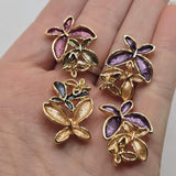 Beautiful multicolor floral 2 Hole Slider Beads featuring three overlapping flowers in rich purple, burgundy, smoky champagne, warm peach, and charcoal blue enamel. Each decorative jewelry focal bead showcases dimensional petals with bright reflective gold outlines and delicate openwork gold flower accents. This coordinated assortment is perfect for handmade bracelets, necklace designs, bead weaving, and multi-strand jewelry.