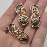 Elegant gold botanical jewelry making beads featuring connected oval shapes with graceful curved branches and softly textured enamel leaves. The charcoal gray sections contrast beautifully with champagne and ivory accents, while polished bright gold outlines add warmth and reflective shine. These unique 2 Hole Slider Beads are ideal for bracelet making, necklace designs, bead weaving, and creative handmade jewelry.