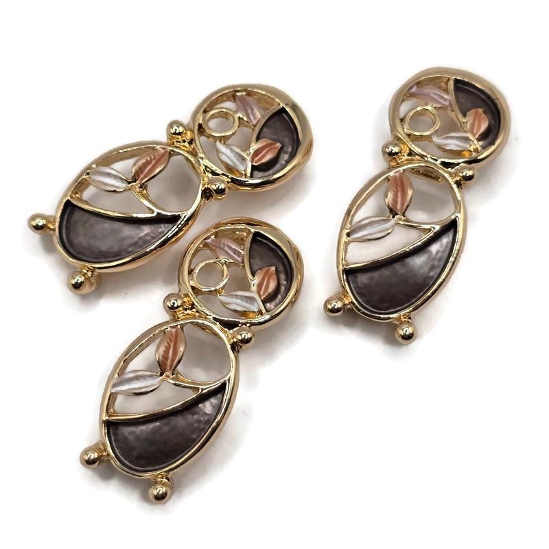 Beautiful bright gold 2 Hole Slider Beads featuring an elongated abstract floral design with two connected oval shapes, flowing gold branches, and delicate enamel leaf accents. Contrasting charcoal gray, champagne, and pearly ivory colors create an elegant neutral combination. These decorative jewelry focal beads feature openwork detailing and highly reflective gold metalwork.