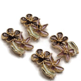 Beautiful bright gold floral 2 Hole Slider Beads featuring two overlapping five-petal flowers in smoky chocolate brown and soft dusty mauve enamel. Each decorative jewelry focal bead includes dimensional gold flower centers, polished metallic outlines, and a delicate pearly ivory leaf accent. These elegant jewelry making components are perfect for handmade bracelets, necklace designs, bead weaving, and multi-strand jewelry.