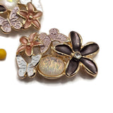 Close-up of decorative floral butterfly slider beads in a polished bright gold finish. Each bead features a large dark brown five-petal flower with a sparkling rhinestone center, smaller peach-colored flowers, and detailed white and lavender enamel butterflies. A luminous pearl-like iridescent accent adds soft flashes of pastel color. Beautiful 2 Hole Slider Beads for double-strand bracelets, necklaces, bead weaving, and feminine handmade jewelry designs.