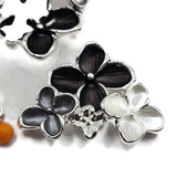 Detailed view of bright silver floral 2 hole slider beads featuring contrasting black, charcoal-gray, and pearl white enamel flowers. The larger black flower has softly textured petals, while the smaller flowers display smoky metallic gray and luminous pearlescent white finishes. Polished silver outlines and raised floral centers create a beautiful monochromatic jewelry-making focal bead.