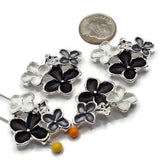 Bright silver floral 2 hole slider bead threaded onto two jewelry-making pins to demonstrate the side-drilled holes and approximately 11 mm strand spacing. The dimensional focal bead features black, smoky gray, and pearl white enamel flowers with polished silver outlines. Decorative multi-strand jewelry component for double-strand bracelets, bead weaving, necklaces, and handmade jewelry designs.