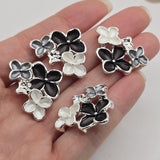Close-up of black, gray, and pearl white enamel flower slider beads in a polished bright silver finish. Each jewelry-making focal bead features one larger black flower with textured petals, a smaller smoky gray flower, and a luminous pearl white flower. Raised silver centers and polished metallic outlines highlight the dimensional floral arrangement, creating elegant decorative components for handmade jewelry.